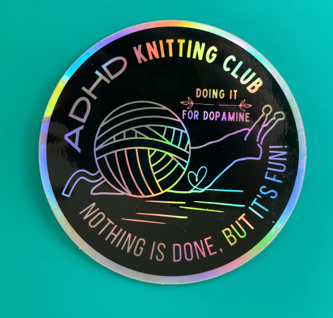Holographic sticker with 'ADHD Knitting Club' text and snail with yarn ball graphic on a teal background
