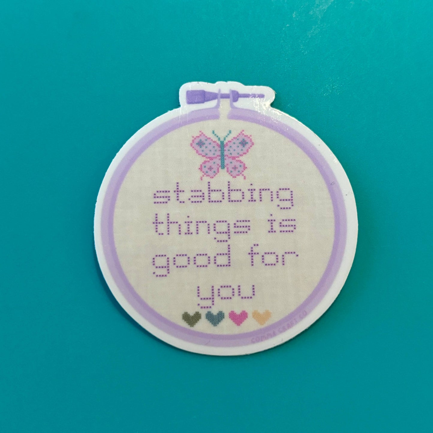 Embroidery hoop with cross-stitched butterfly and stabbing things is good for you text on a teal background