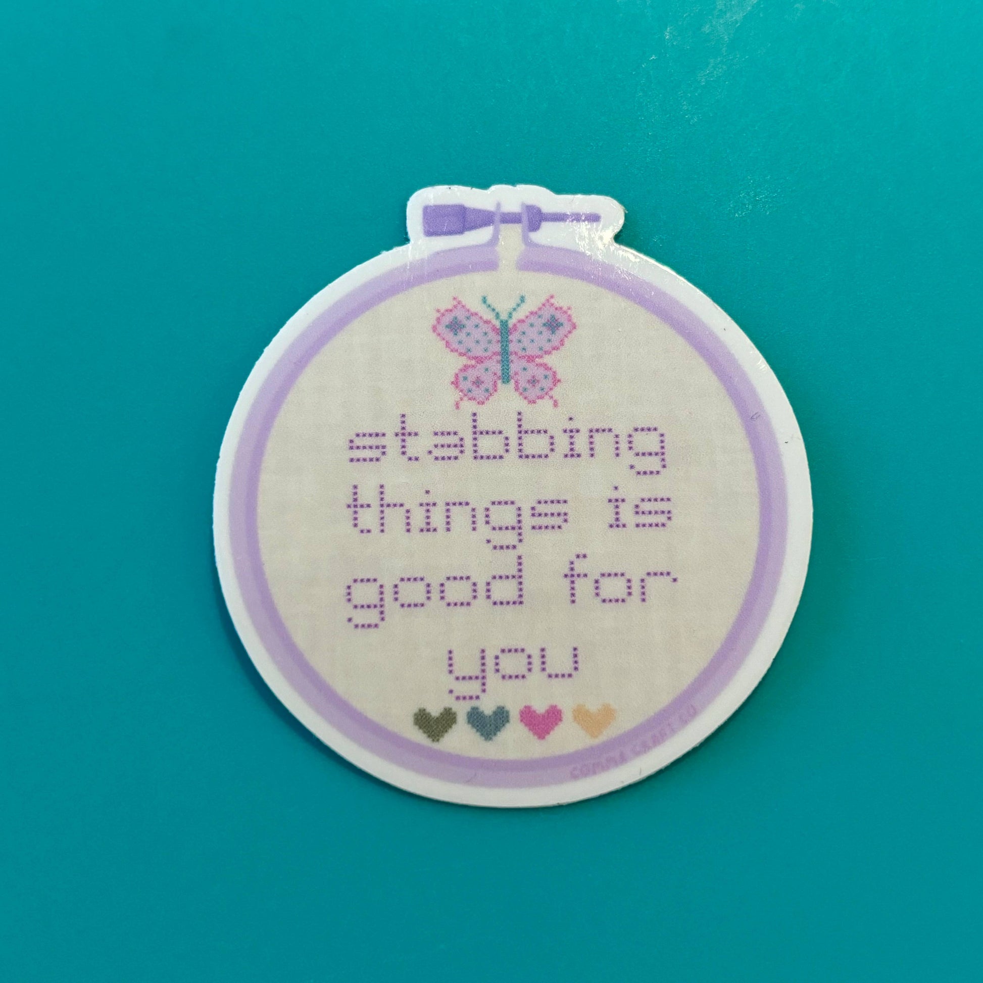 Embroidery hoop with cross-stitched butterfly and stabbing things is good for you text on a teal background